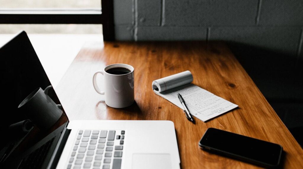 A cozy workspace with a laptop, coffee, notebook, and smartphone on a wooden table.