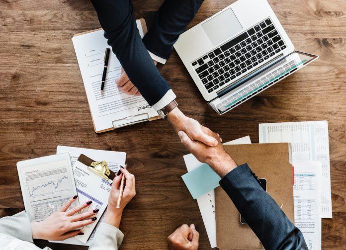 Two people shaking hands over a business meeting with documents and a laptop.