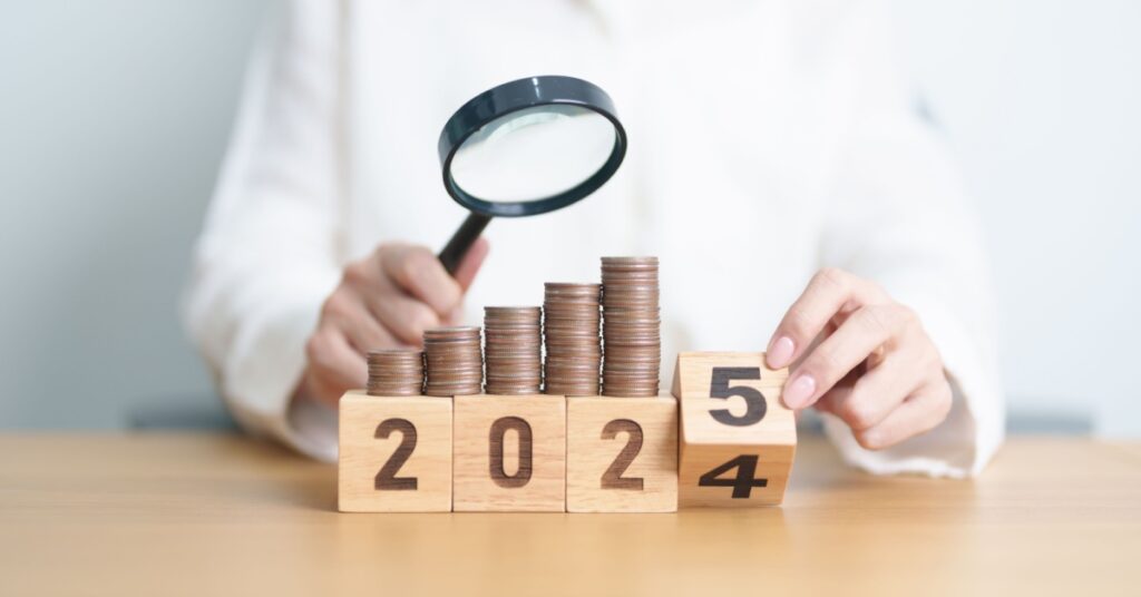 Magnifying glass over stacked coins and wooden blocks showing year change from 2024 to 2025.